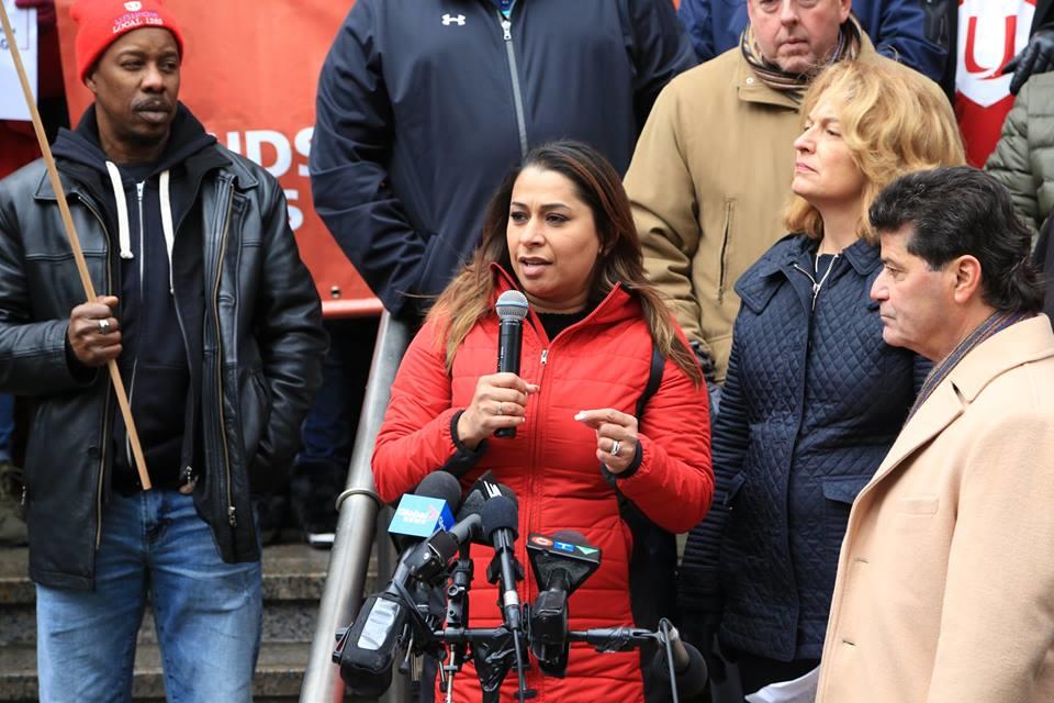 Pre-pandemic photograph of Naureen Rizvi speaking at a microphone at a rally for workers’ rights.