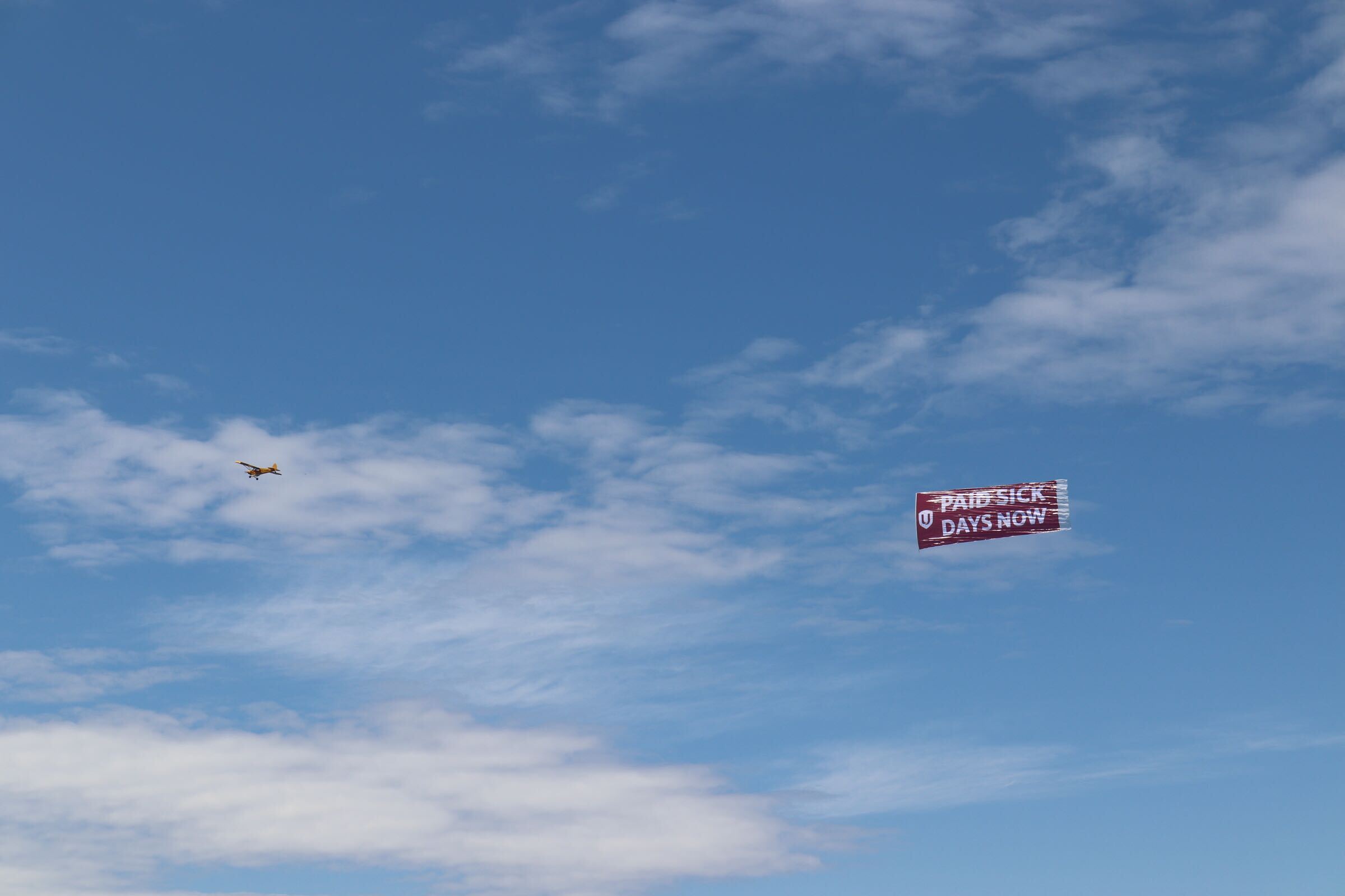 An airplane flys, pulling a banner that reads, "Paid Sick Days Now."