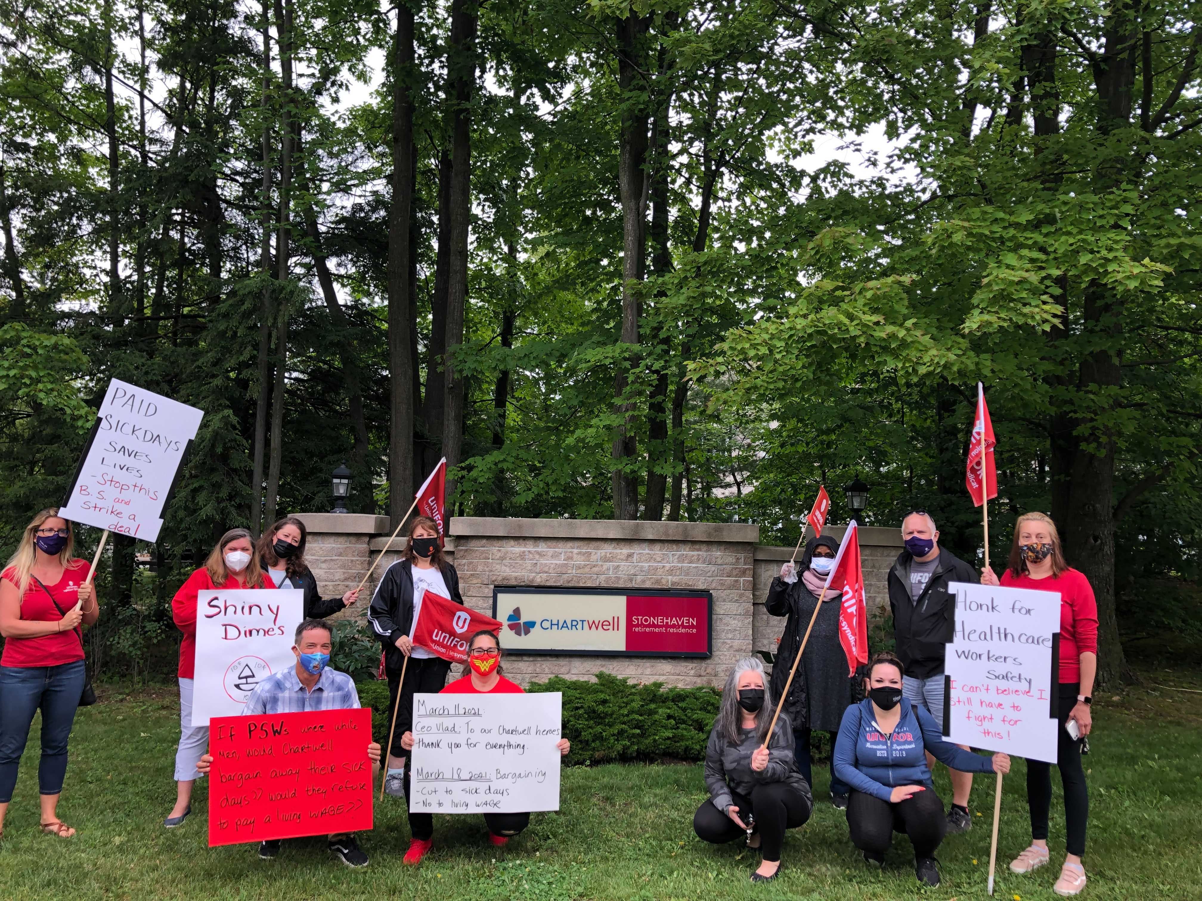 Health care workers at a rally in front of a Chartwell's sign outside on a decrative wall. Workers are holding signs and flages posing.
