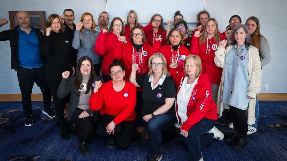 A group of more than 20 Unifor members, meany wearing red, post for photo with serious faves while holding fists in the air.