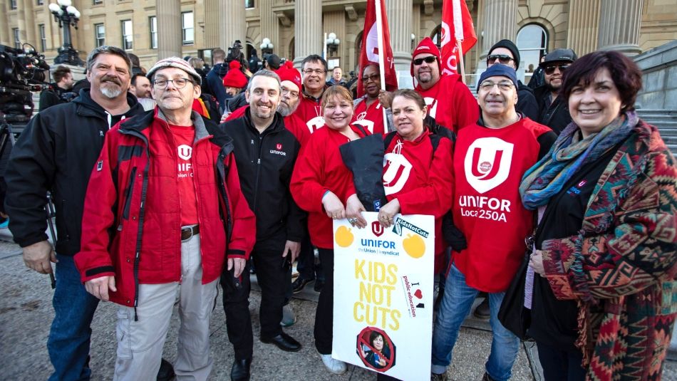 Unifor members posing in front of the Alberta legislature holding flags and a sign reading 'Kids not cuts'