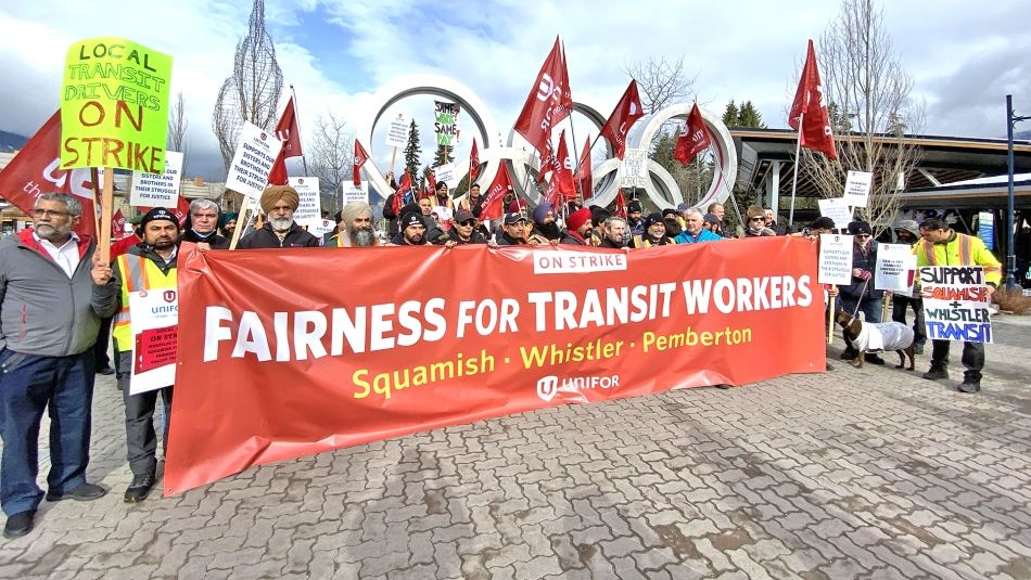 Large group of rally participants with flags and placards posing with a "Fairness for Transit Workers" banner in front of Whistler Village's Olympic rings..