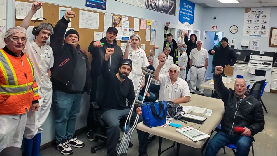 « Dix-huit membres d’Unifor posent pour la photo en levant leurs poings dans les airs dans une salle à manger. Il y en a parmi eux qui portent un uniforme blanc et un filet à cheveux. »