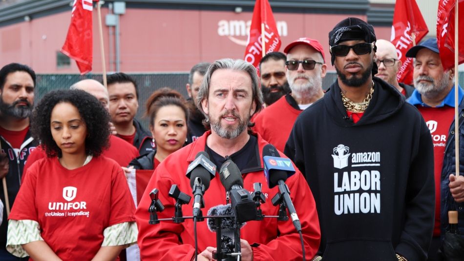 Gavin McGarrigle parle au micro devant des travailleuses et travailleurs portant des chandails rouges et une installation d’Amazon en arrière-plan.