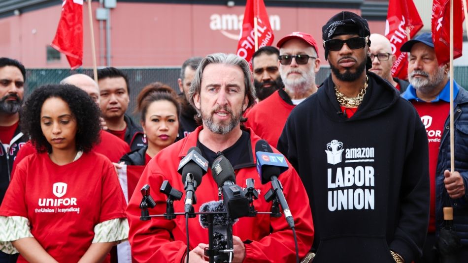 Gavin McGarrigle speaking in front of several microphones in front of a group of Unifor members wearing red shirts."