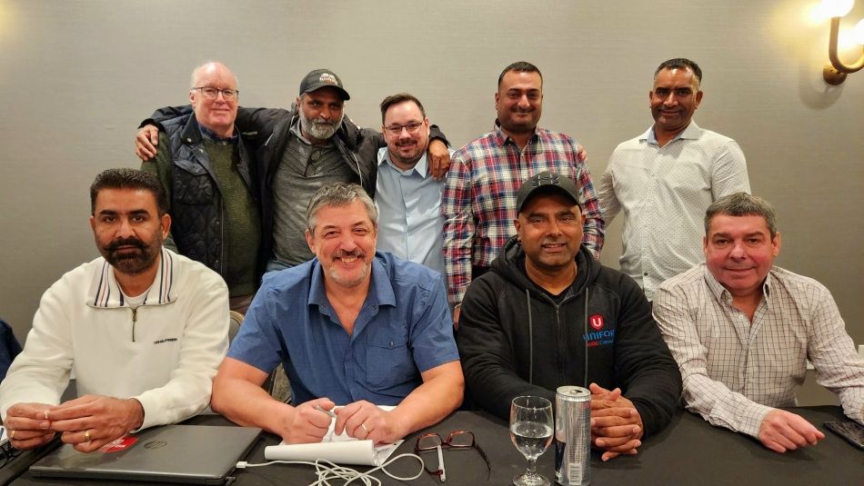 Four men sitting at a table and five others standing behind facing forward smiling.