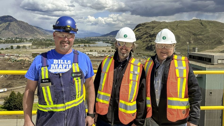 Trois personnes portant des casques de protection et des vêtements à haute visibilité en plein air sur le toit d'une usine de pâte à papier avec des montagnes en arrière-plan. 