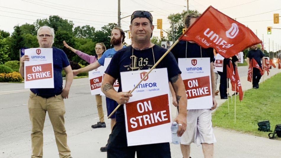 Des personnes se tiennent sur un piquet de grève avec des pancartes portant l'inscription "On Strike". L'homme au premier plan tient un drapeau d'Unifor.
