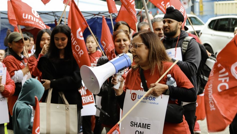 Une femme parle dans un mégaphone et des travailleurs tenant des drapeaux se tiennent derrière elle.