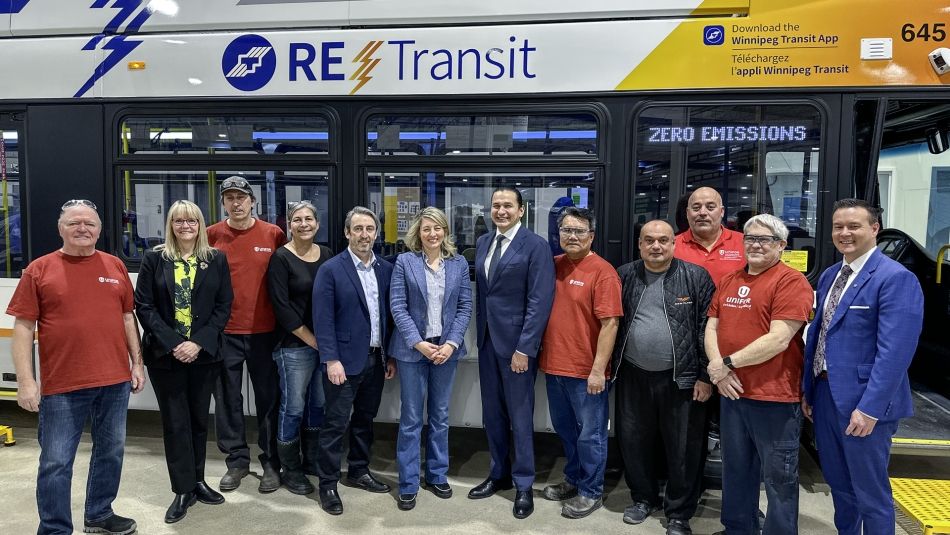 A dozen people post for a photo standing beside a bus inside a warehouse.