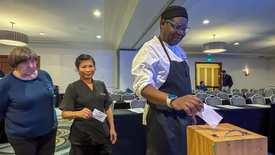 One worker puts paper into a ballot box while two are lined up behind him.