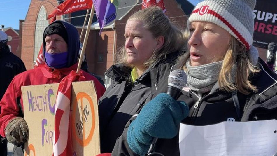 A woman, wearing a Unifor toque and turquoise mitts, speaking into a microphone standing beside a man and woman looking to their left, holding signs and flags.