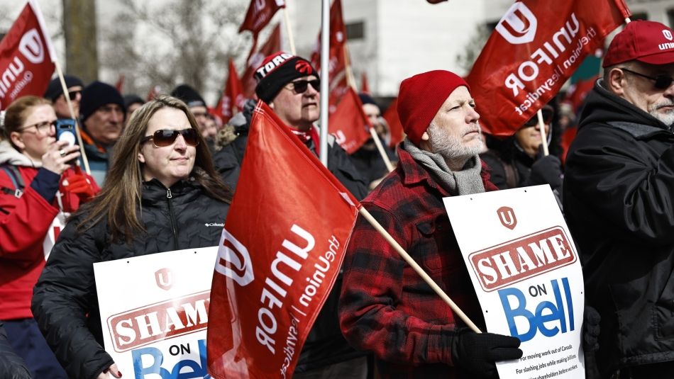 A man and a woman stand with Shame on Bell signs with a crowd behind them