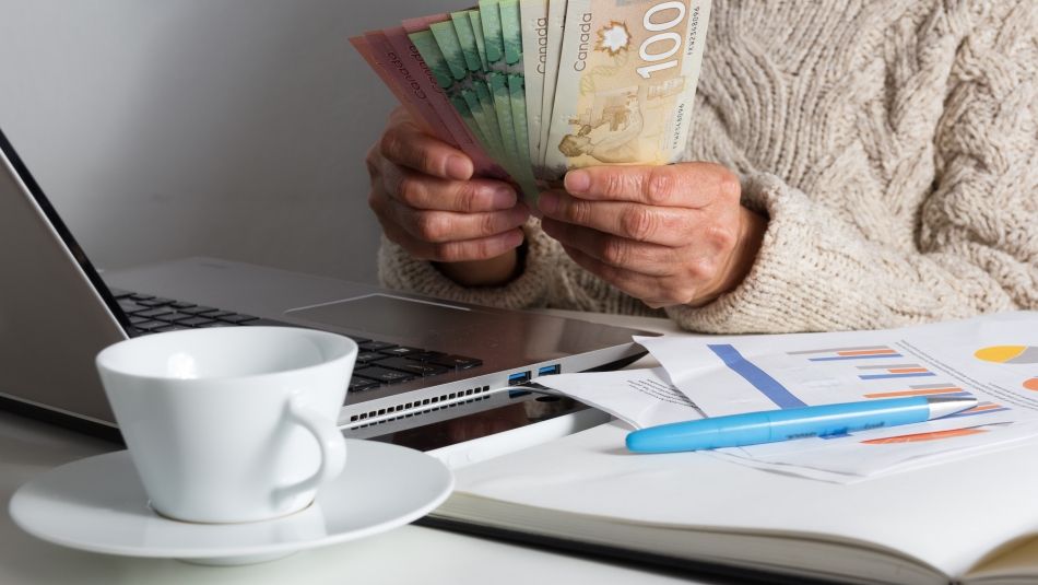 une personne assise devant un ordinateur portable à côté d'un cahier et d'une tasse de thé tenant de l'argent différents billets disposés en éventail