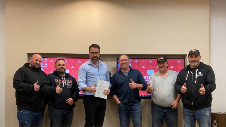 Six people giving a thumbs up standing in front of two presentation screens