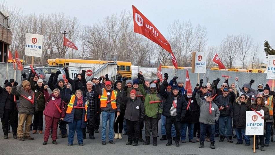 A group of people, raising their firsts in the air and holding white Unifor placards in front of a chain-link fence with yellow school buses in the background.