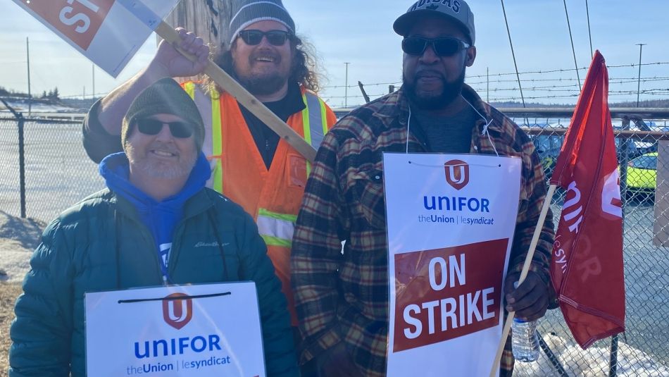 Three Unifor members hold strike signs and flags as they stand on the picket line