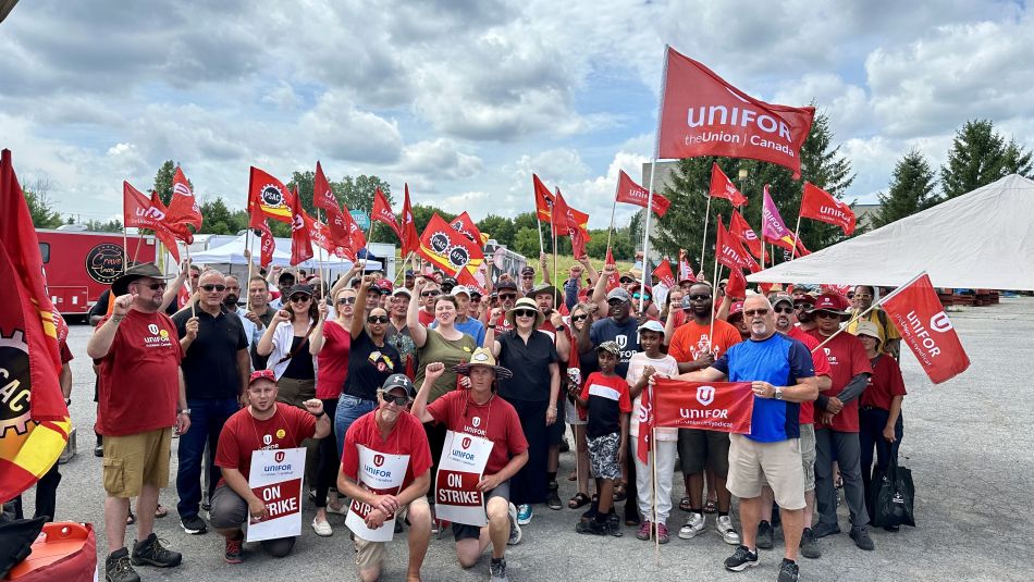 A large group of union members and supporters gather outdoors, raising fists and waving red Unifor and PSAC flags. Several people in front hold “On Strike” signs.