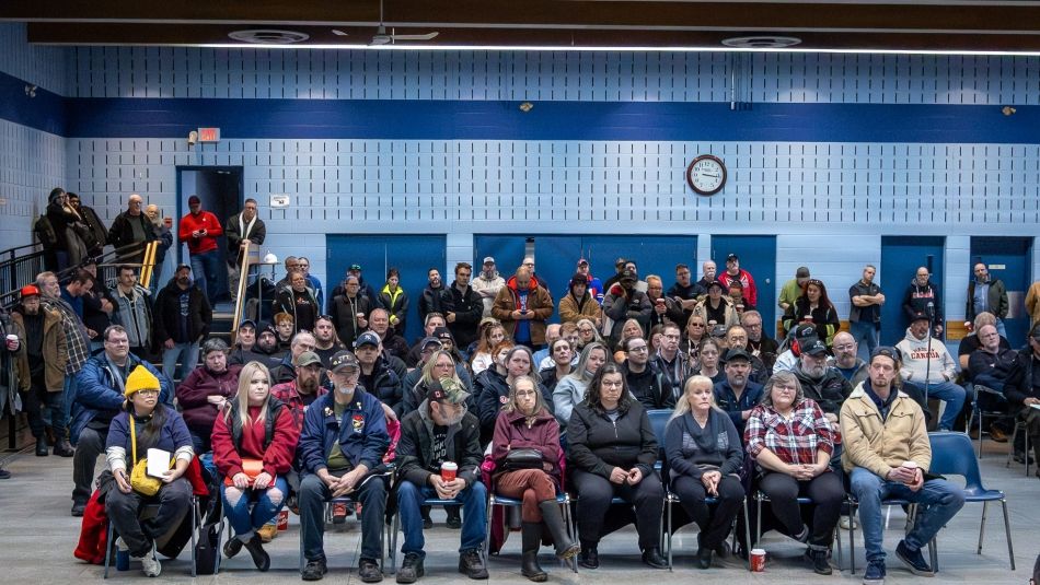 A large group of people sit facing forward in rows of chairs in a community hall, listening attentively; a clock and blue walls are visible in the background.