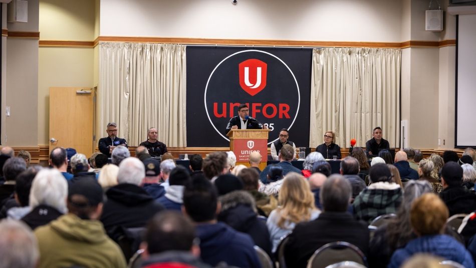  A speaker addresses a seated audience from a podium on stage at a Unifor union event, with several panelists seated behind them. 