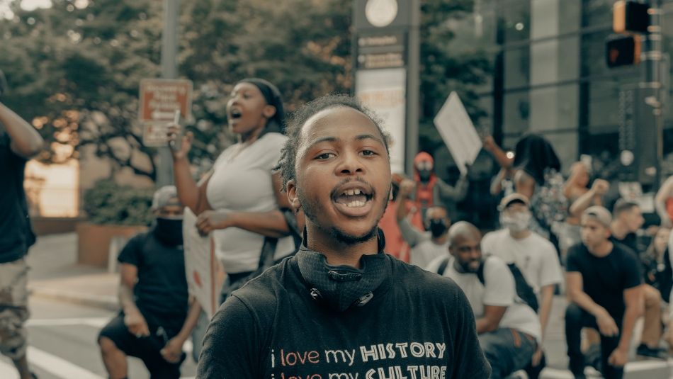 Black youth vocalizing in the street with protesters in the background. He is wearing a black t-shirt that says, “I love my history, I love my culture, I love my people, I love me.”