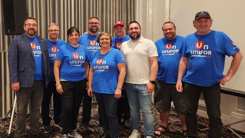 A group of people wearing blue Unifor Quebec shirts pose in front of a curtain and speaker.