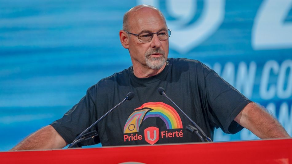 Unifor National Secretary-Treasurer Len Poirier wearing a Unifor Pride shirt, speaking at a podium.