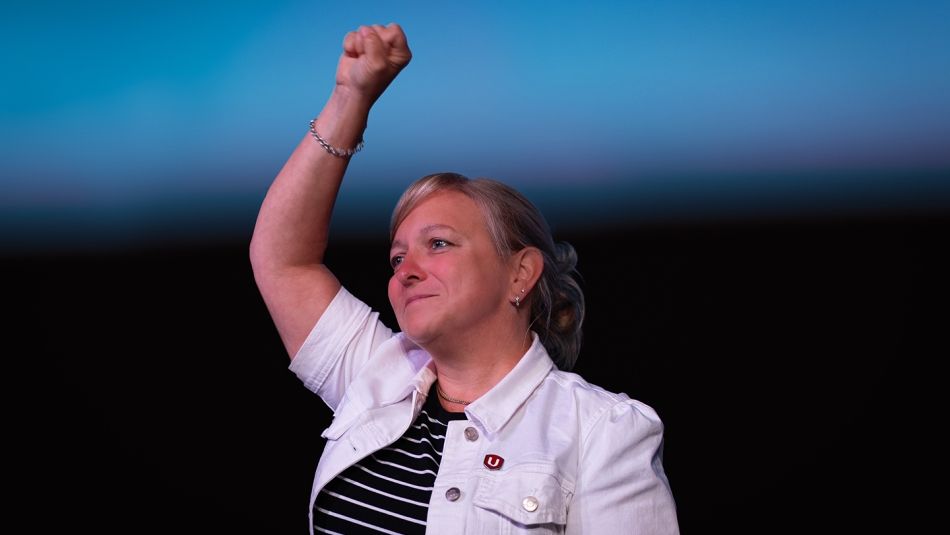 A blonde woman wearing a white jacket raises her right fist in the air.