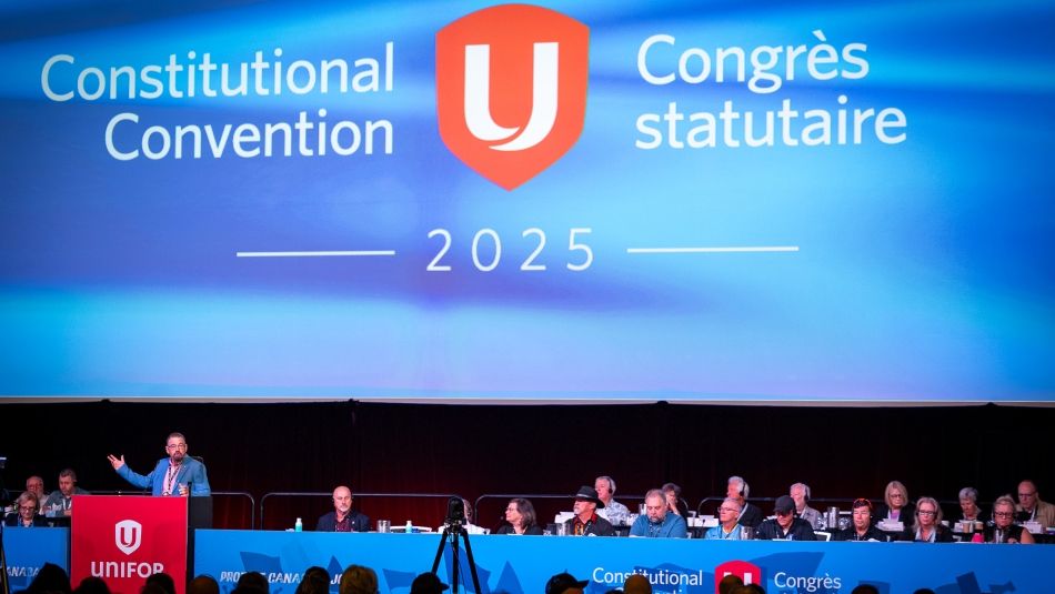 A large Unifor stage with people sitting and blue backdrop featuring Unifor shield logo.