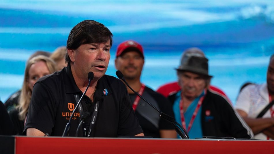 A man wearing a black Unifor shirt speaks at a podium with a crowd behind him.