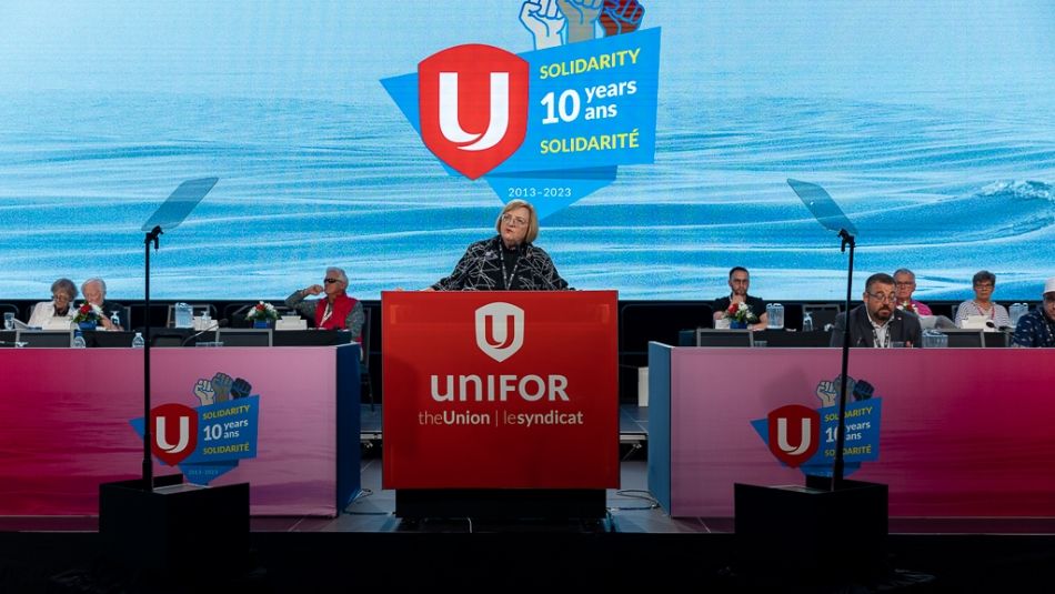 Lana Payne stands at the podium on stage with the Unifor graphic in the background.