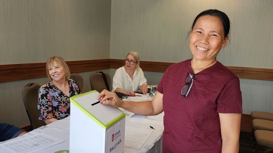 Une femme souriante dépose un bulletin de vote dans une urne tandis que d'autres personnes sont assises à une table à proximité.