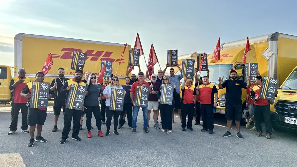 A group of people holding signs and flags stands in front of DHL-branded trucks, in a union rally
