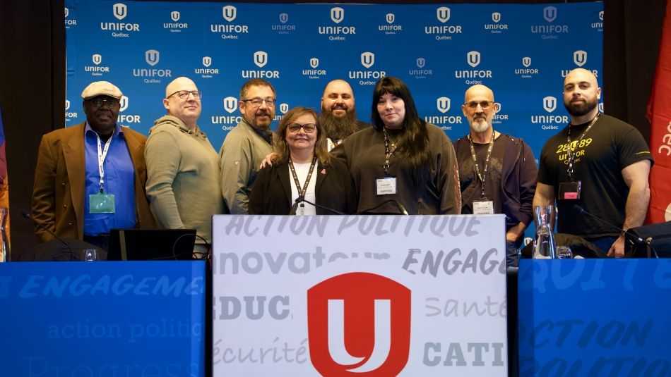Eight Quebec Council Executive members posing behind a podium in Unifor decal and in front of a blue Unifor banner