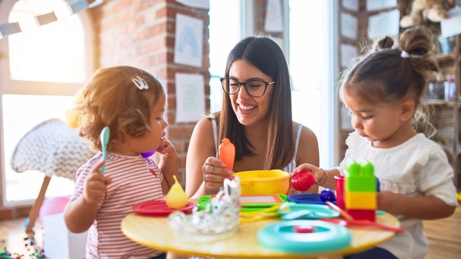 An early childhood educator sits at a table with two young children.