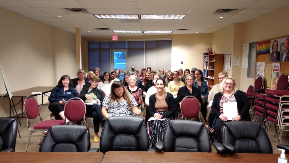  A group of people sit n a room, with four empty black chairs in the front row.