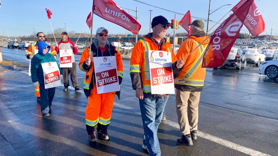Unifor Local 100 Autoport Workers United in Strike Action A group of workers from Unifor Local 100, identifiable by their signs and flags, are participating in a strike. They are wearing high-visibility safety gear and are marching in a parking area under clear skies, conveying a sense of solidarity and purpose.