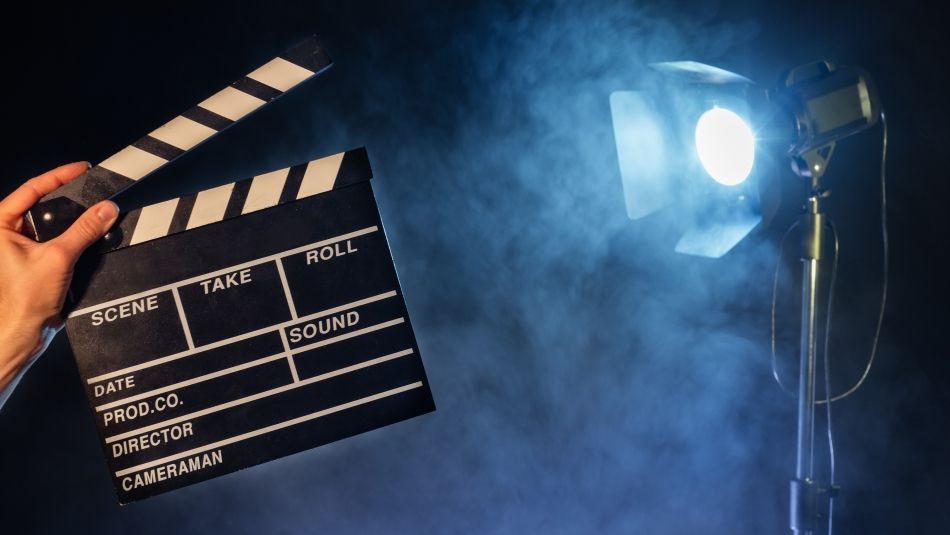 A hand holds a clapperboard against a smoky backdrop, with a bright studio light illuminating the scene.