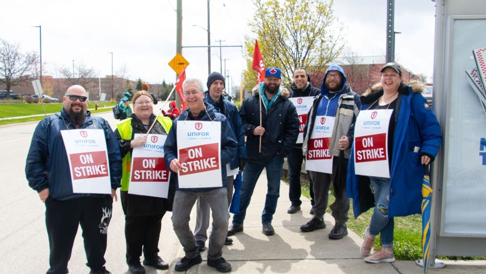 “Group of picketers wearing On Strike placards and smiling outside at a bus stop.”
