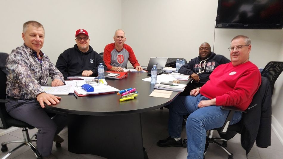 Five men sitting at a table with a flatscreen TV in the wall behind them to the right.