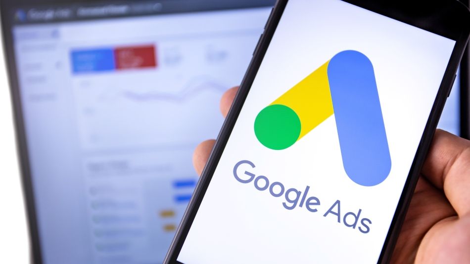 A hand holding a phone that has Google Ads on the screen with a green dot, and yellow and blue lines. There is a computer screen with a line graph in the background.