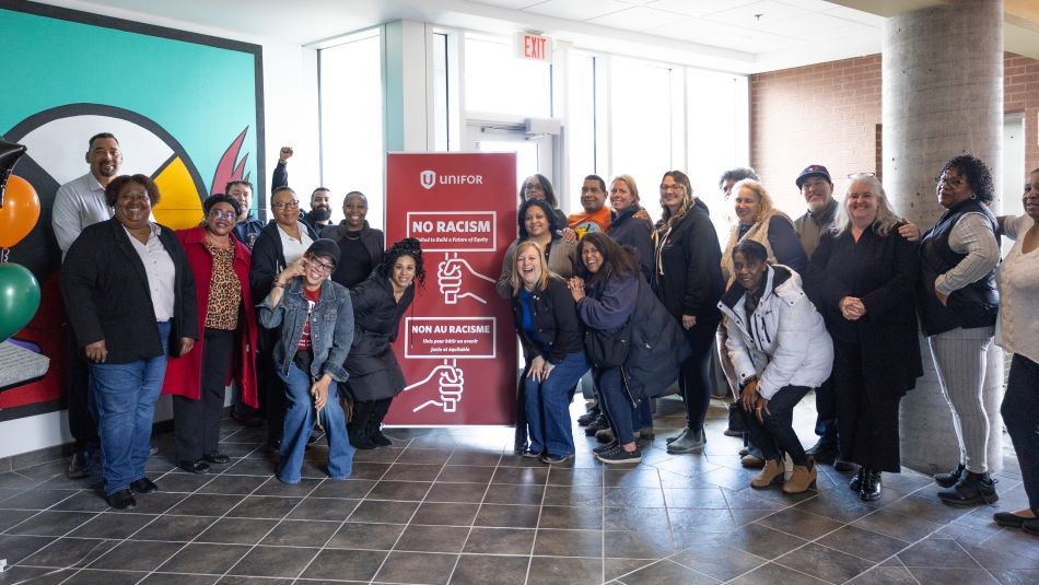 A large group on either side of a red Unifor banner, a hand holding a sign reads, no racism. United to build of future of equity