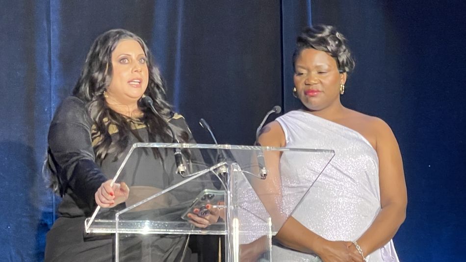 A woman speaks at a podium, while another woman in a white dress stands next to her.