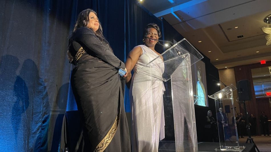 Two women stand at a podium with a blue curtain background.