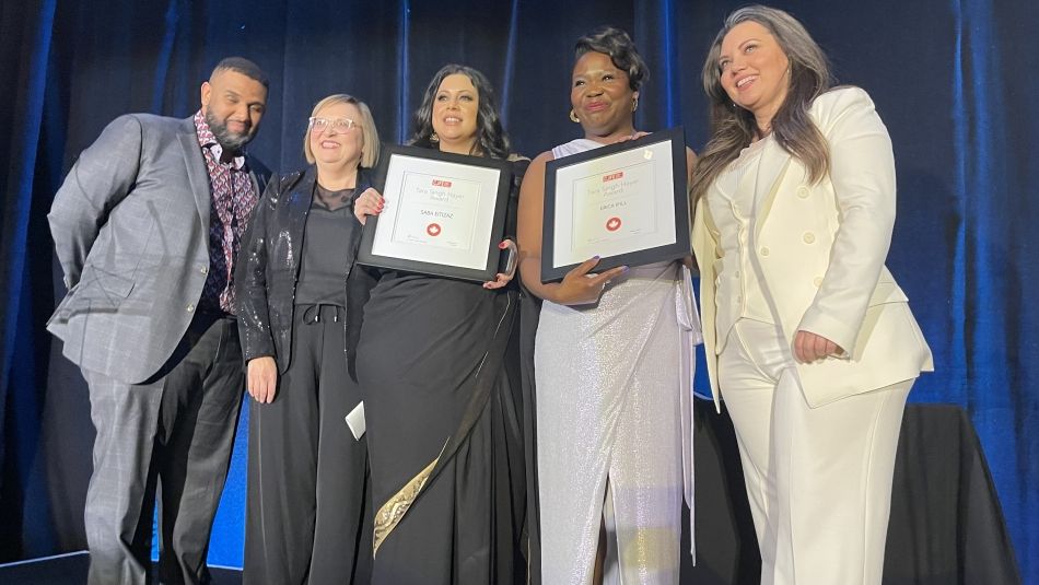 Two women hold up awards, surrounded by Lana Payne and two hosts on a blue curtained stage