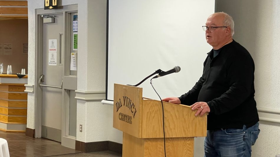 A man wearing jeans and a dark top and glasses with grey hair speaks into a microphone at a podium. A white projection screen is in the background.