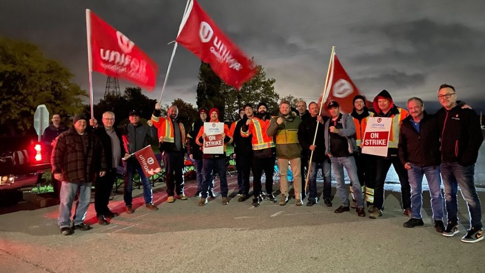 A large group of workers stand outside at midnight holding strike signs and flags