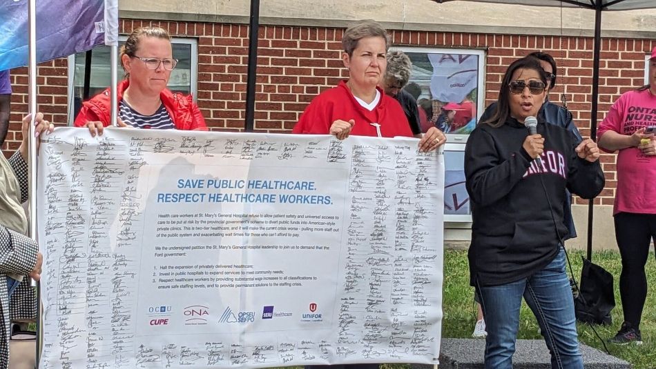 Two men holding up a banner petition signed by Ontario Health Care workers. A women speaks into a mic.