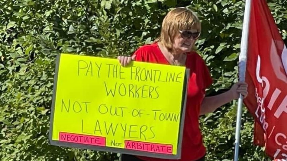 A women marching along a road holding up a neon sign and red Unifor flag.