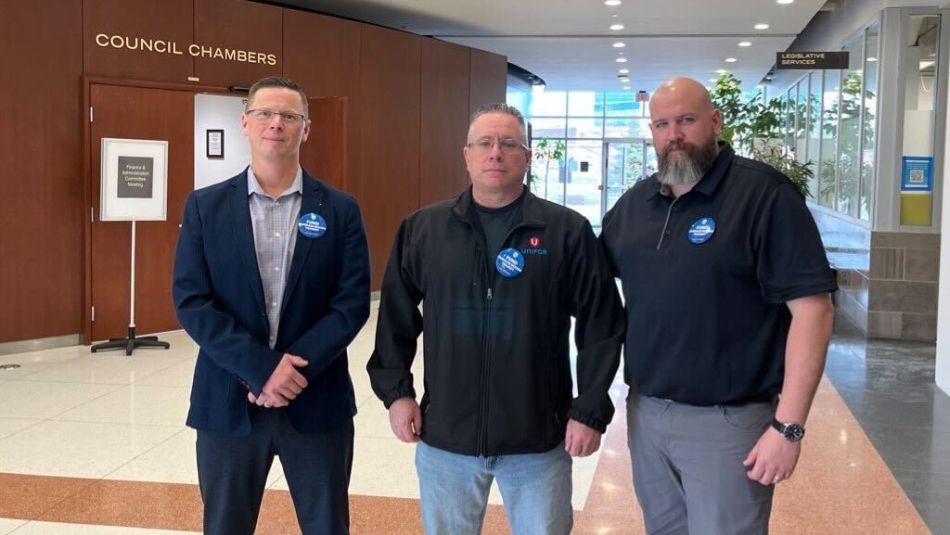Three men wearing blue buttons standing in front of council chambers.